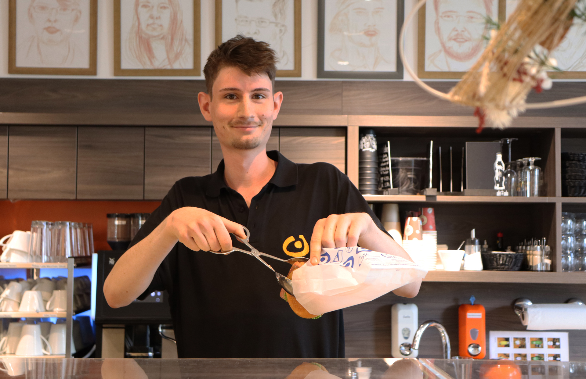 Mann im Restaurant schneidet mit einer Schere eine Zutat für ein Gericht. Im Hintergrund sind Küchenutensilien und Speisekarten sichtbar.