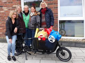 Drei Frauen stehen vor einem Fahrrad. Das Fahrrad hat einen großen Korb. Im Korb sind Spielsachen.