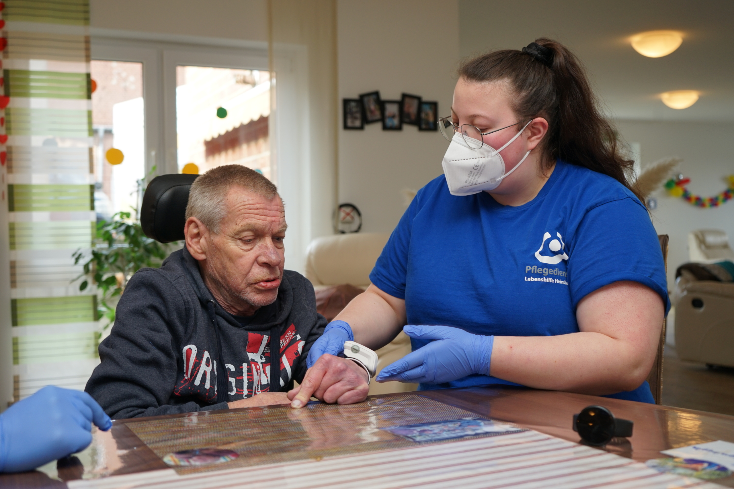 Ein älterer Mann und eine Frau sitzen an einem Tisch. Die Frau hilft dem Mann. Beide tragen Handschuhe.