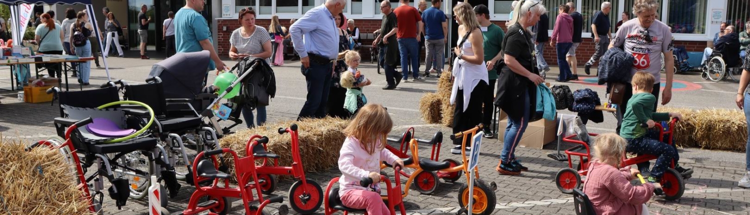 Kinder spielen auf Dreirädern auf einem Fest mit vielen Besuchern im Hintergrund. Bunte Fahnen wehen, und es herrscht eine fröhliche Atmosphäre.