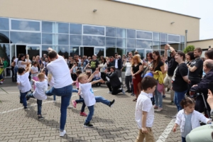 Kinder spielen draußen vor einem Gebäude. Manche Kinder hüpfen und lachen. Es sind viele Menschen im Hintergrund zu sehen.