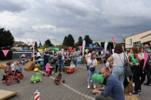Familienfest mit Kindern, die auf Spielzeugfahrzeugen spielen, umgeben von bunten Ständen und Besuchern. Wolkiger Himmel im Hintergrund.