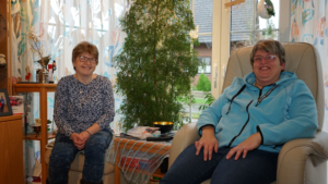 Zwei Frauen sitzen in einem hellen Raum mit Pflanzen, entspannt und in geselliger Atmosphäre.