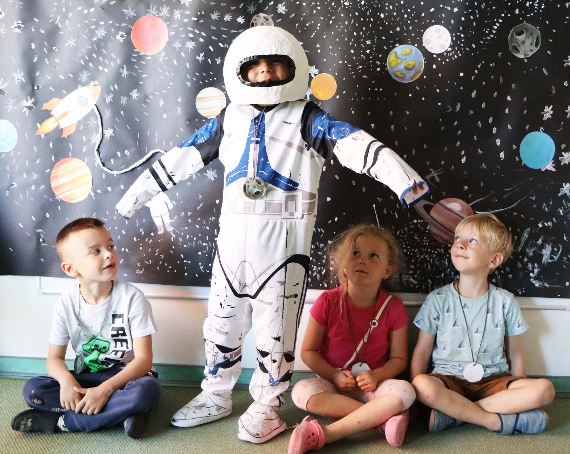 Kinder sitzen und stehen vor einer Wand mit Sternen. Ein Kind trägt einen Raumanzug.