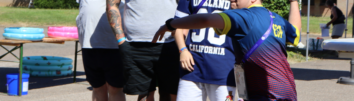 Junger Mann in Sportkleidung wirft einen Ball, während eine Gruppe von Zuschauern im Hintergrund zusieht. Der Ort scheint ein Freiluft-Event oder ein Sportfest zu sein, umgeben von Zelten und Bäumen.
