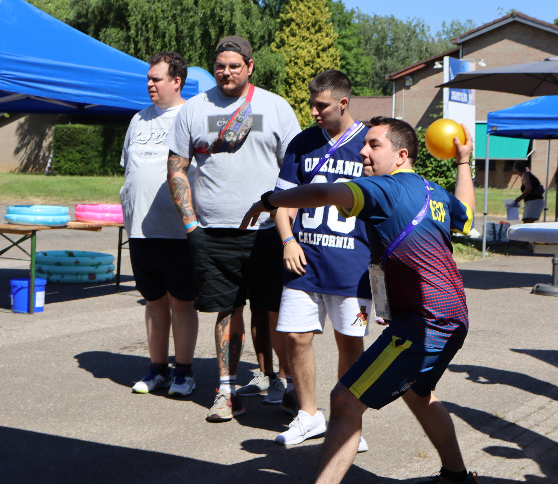 Junger Mann in Sportkleidung wirft einen Ball, während eine Gruppe von Zuschauern im Hintergrund zusieht. Der Ort scheint ein Freiluft-Event oder ein Sportfest zu sein, umgeben von Zelten und Bäumen.
