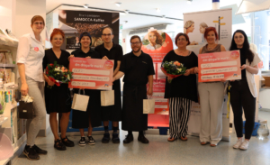 Gruppe von sechs Frauen, die bei einer Veranstaltung in einem Einkaufszentrum Blumensträuße halten, umgeben von Werbebannern. Sie zeigen ihre Unterstützung für eine lokale Initiative.