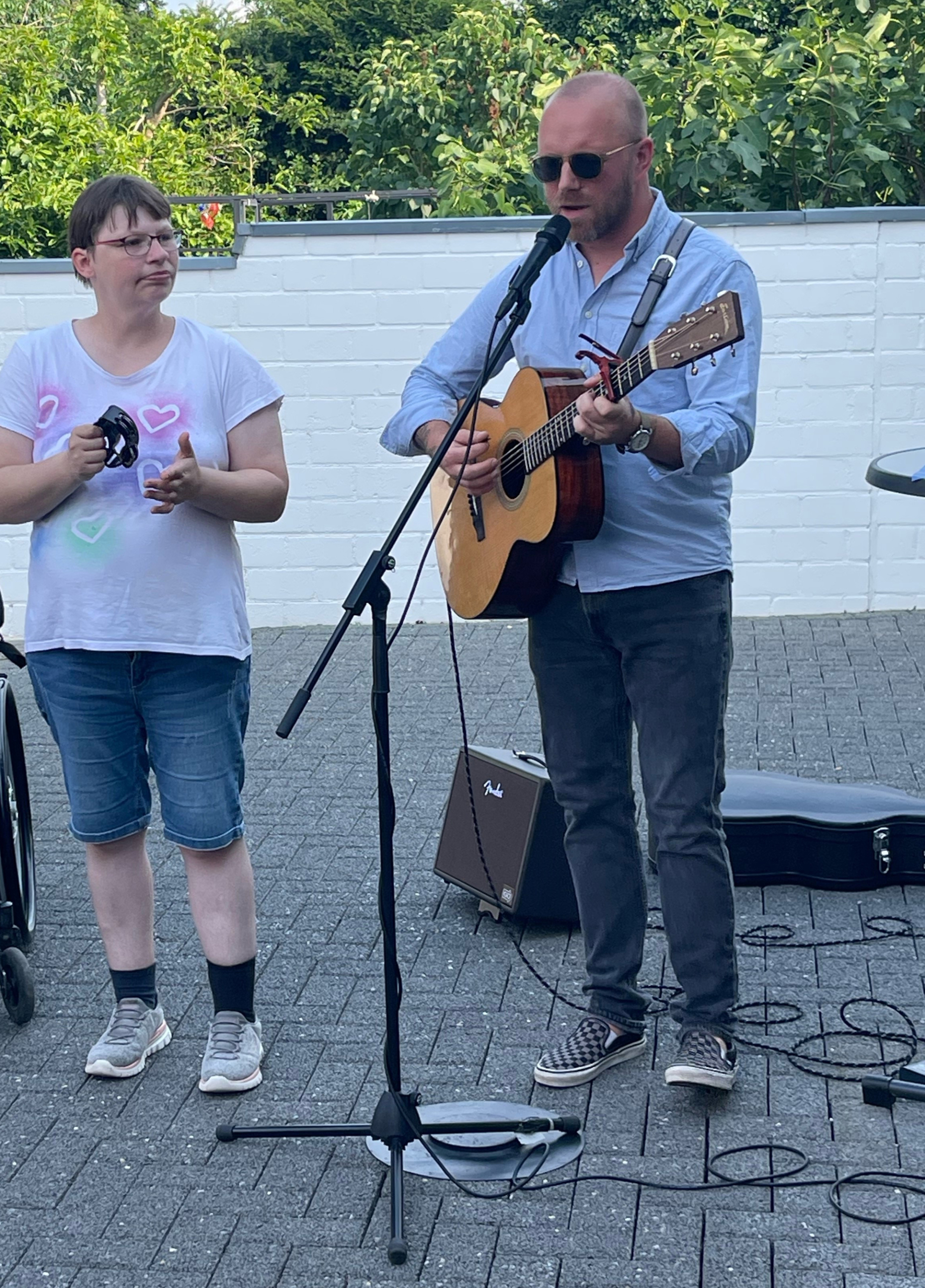 Eine Frau steht neben einem Mann, der Gitarre spielt und singt, während sie ein Mikrofon hält. Die Szene findet im Freien statt, umgeben von einer entspannten Atmosphäre.