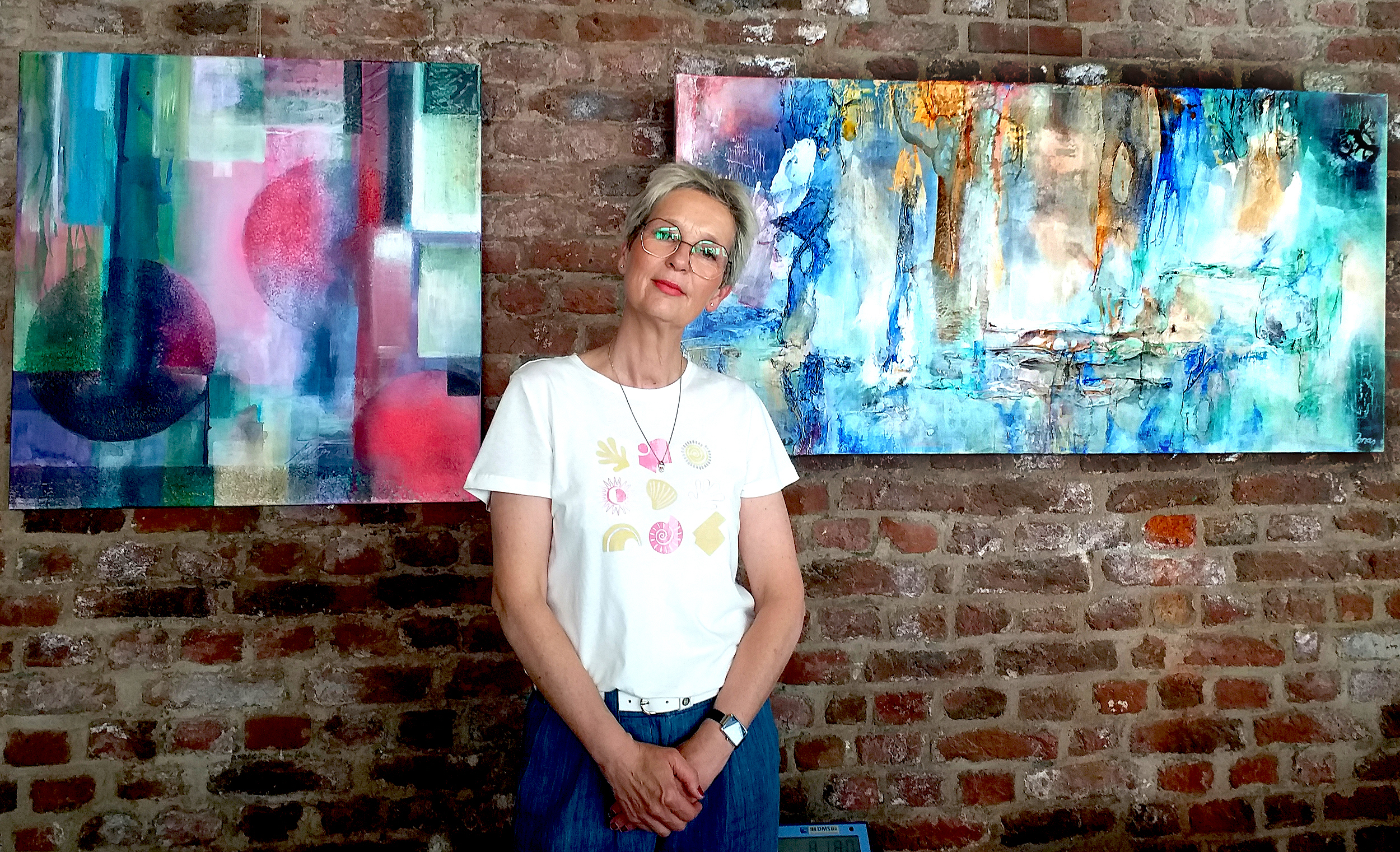 Eine Frau steht vor einem Bild. Das Bild hat bunte Farben und sieht aus wie ein Landschaftsbild. Die Frau trägt ein weißes T-Shirt.