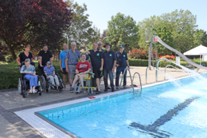 Eine Gruppe Menschen steht vor einem Schwimmbad. Manche sitzen im Rollstuhl.