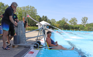 Mann sitzt am Beckenrand eines Schwimmbads und nutzt ein Rudergerät, während andere Personen im Hintergrund schwimmen. Sonniger Tag mit klarem Himmel und Bäumen im Hintergrund.