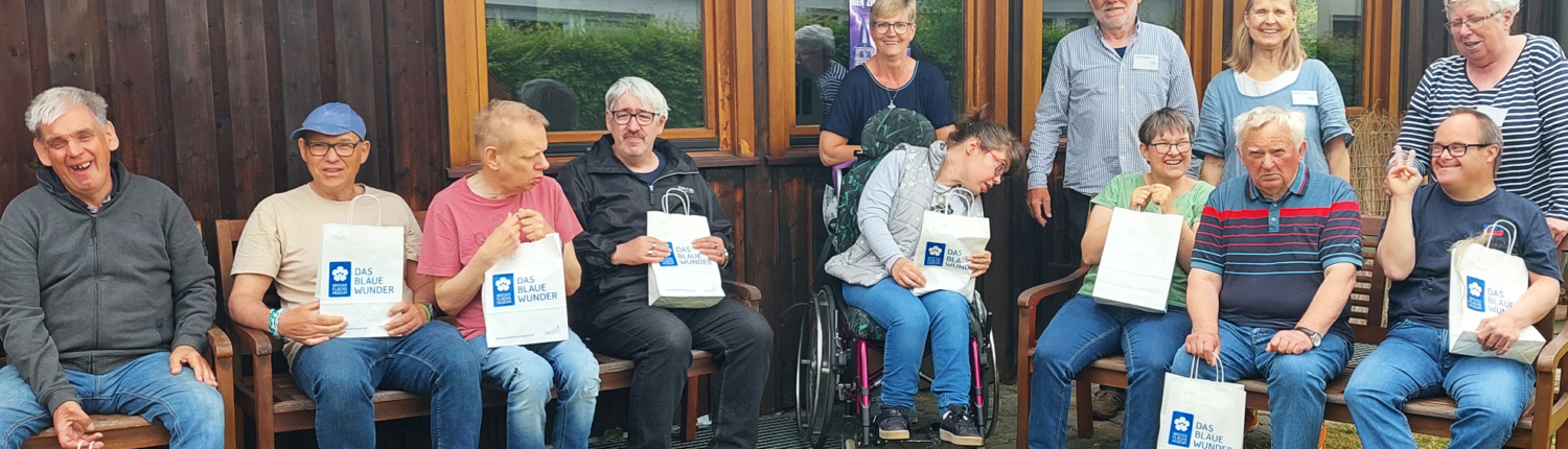 Eine Gruppe älterer Menschen sitzt draußen auf Bänken. Sie halten Papiertüten und lächeln.