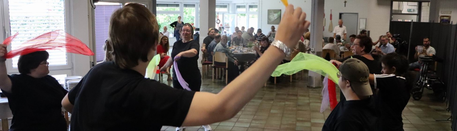 Menschen tanzen mit Tüchern in einem Raum. Manche haben kurze Haare.
