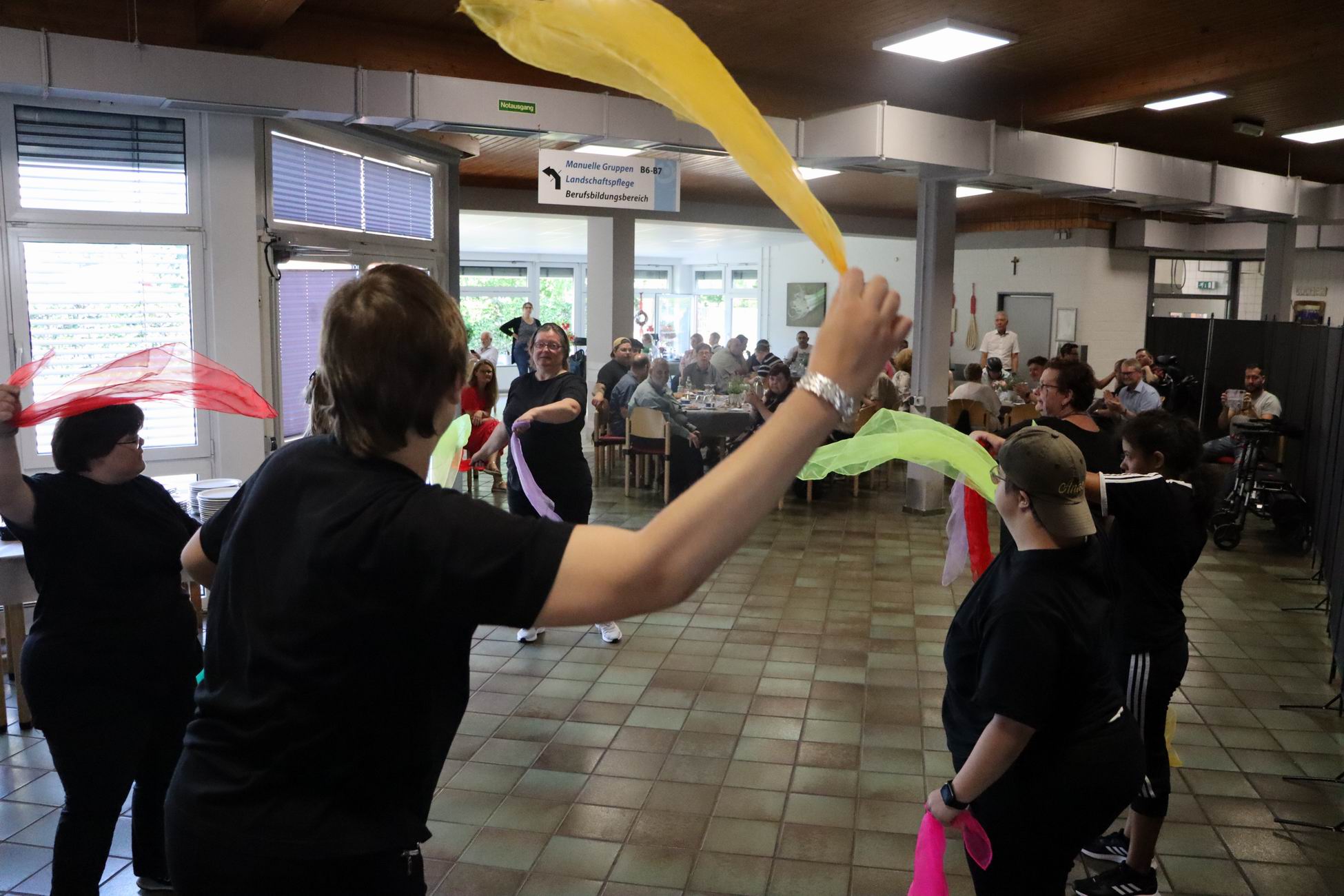 Menschen tanzen mit Tüchern in einem Raum. Manche haben kurze Haare.