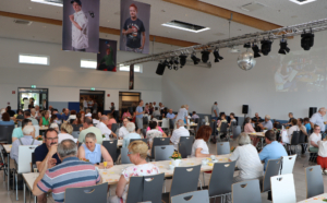 Viele Leute sitzen an Tischen in einem Saal. Es gibt Blumen auf den Tischen.