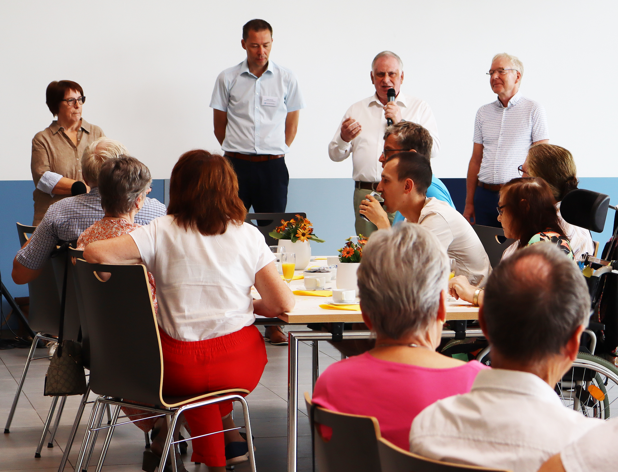 Menschen sitzen um einen Tisch. Ein Mann steht und spricht.