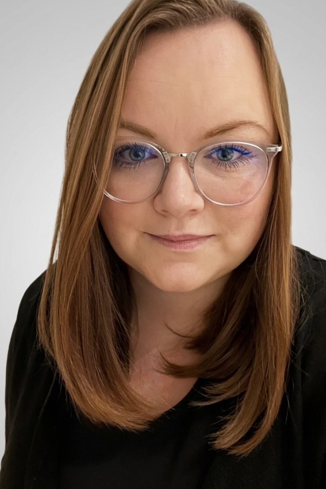 Porträt einer Frau mit langen, welligen Haaren und Brille, die in einem professionellen Umfeld steht. Sie trägt ein schwarzes Oberteil und hat einen freundlichen Gesichtsausdruck.