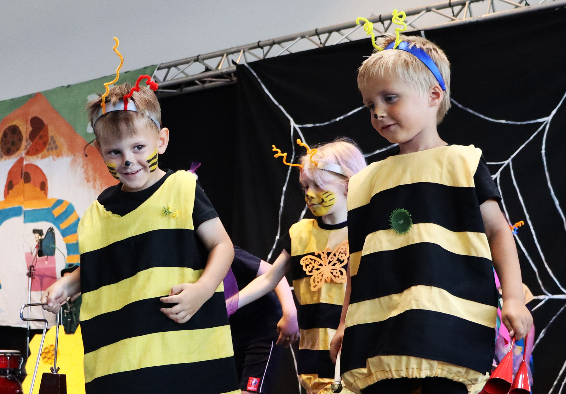 Kinder in Bienenkostümen führen eine Aufführung auf der Bühne auf, umgeben von bunten Requisiten und fröhlicher Atmosphäre.