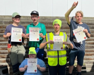 Gruppe von Jugendlichen in Sicherheitskleidung, die stolz ihre Zertifikate halten und dabei lächeln. Hintergrund zeigt eine Baustelle.