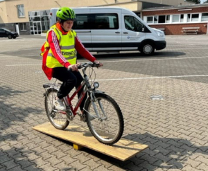 Ein Kind fährt Fahrrad auf einem Brett. Es trägt einen Helm und eine Warnweste.