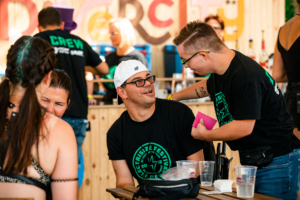 Zwei Männer in schwarzen T-Shirts mit grünem Logo interagieren in einem lebhaften Restaurant, während eine Frau im Hintergrund sitzt. Die Szene vermittelt eine freundliche Atmosphäre und zeigt das Personal bei der Arbeit.