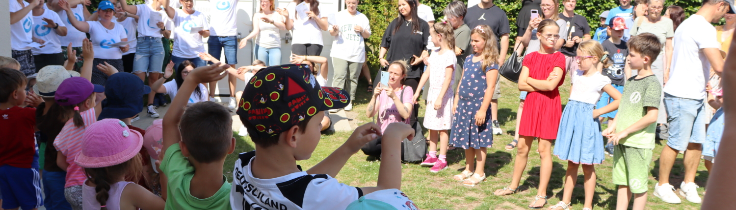 Eine Gruppe von Kindern und Erwachsenen versammelt sich im Freien zu einer Feier oder Veranstaltung. Die Kinder stehen im Vordergrund und zeigen Freude, während die Erwachsenen im Hintergrund beobachten. Es ist ein sonniger Tag mit blauem Himmel und grüner Umgebung.