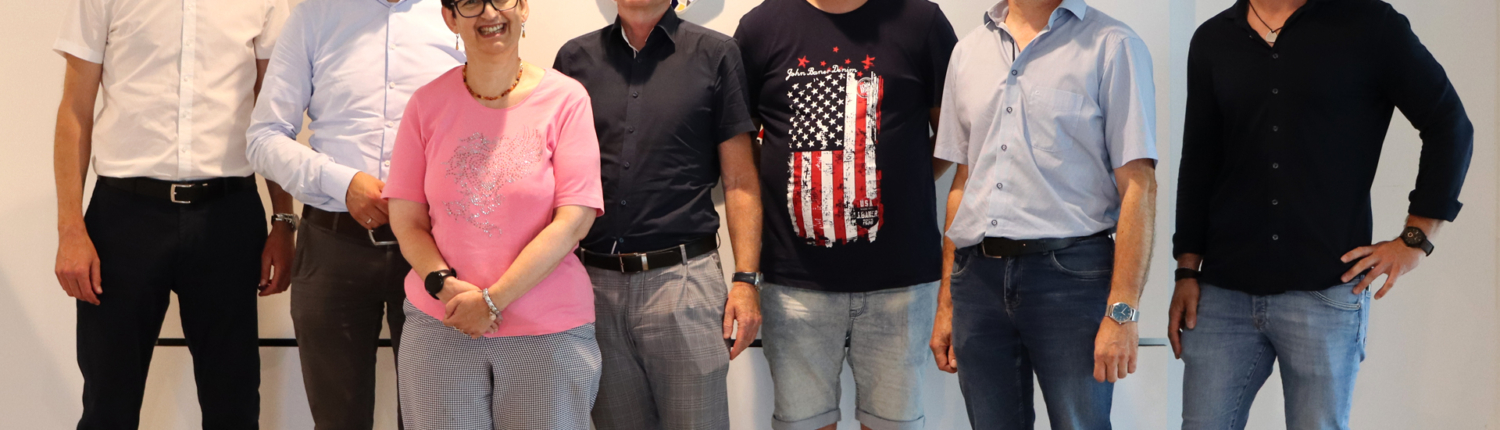 Sechs Männer und eine Frau stehen zusammen vor einer Wand. Sie lächeln.