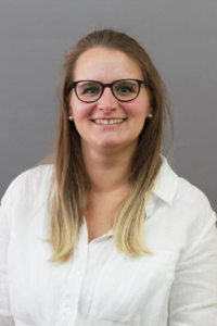 Eine Frau lächelt. Sie trägt eine weiße Bluse und eine Brille.