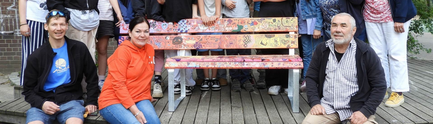 Gruppe von Jugendlichen und Erwachsenen versammelt im Freien, lächelnd vor einer Holzbank. Die Szene zeigt eine freundliche Gemeinschaft, die an einem sozialen oder schulischen Projekt beteiligt ist.