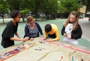 Gruppenaktivität im Freien, bei der vier Personen gemeinsam an einem Tisch arbeiten und kreative Bastelprojekte durchführen. Im Hintergrund ist eine grüne Umgebung sichtbar, die eine entspannte Atmosphäre schafft.