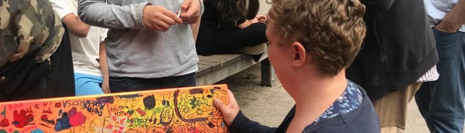 Eine Gruppe von Menschen, darunter ein Mann im Anzug und eine Frau mit lockigen Haaren, arbeitet gemeinsam an einer bunten, bemalten Bank im Freien. Kinder und Erwachsene beobachten interessiert.
