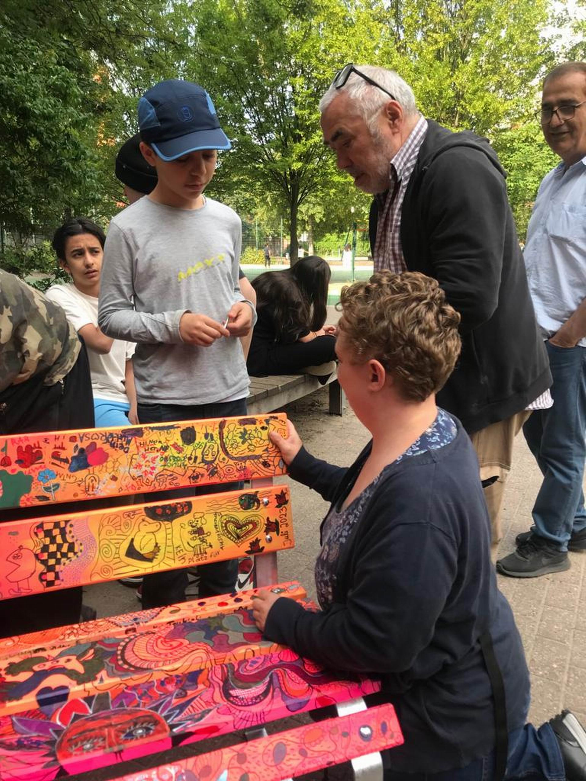 Eine Gruppe von Menschen, darunter ein Mann im Anzug und eine Frau mit lockigen Haaren, arbeitet gemeinsam an einer bunten, bemalten Bank im Freien. Kinder und Erwachsene beobachten interessiert.