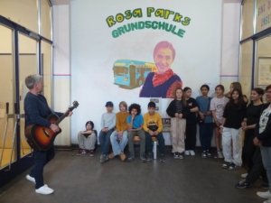 Ein Mann spielt Gitarre. Kinder sitzen und schauen ihm zu. Sie sind in einem Raum mit einem großen Wandbild.