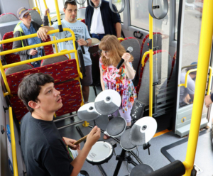 Ein Musiker spielt Schlagzeug in einem Bus, während Passagiere aufmerksam zuschauen. Die Szene zeigt eine lebendige Atmosphäre und die Interaktion zwischen Kunst und öffentlichem Verkehr.
