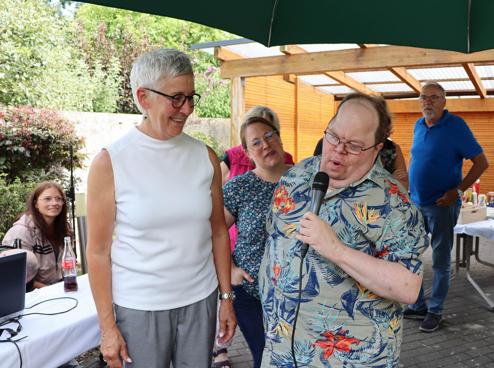 Ein Mann spricht ins Mikrofon. Eine Frau steht daneben. Sie lächeln. Es sind noch andere Leute da.