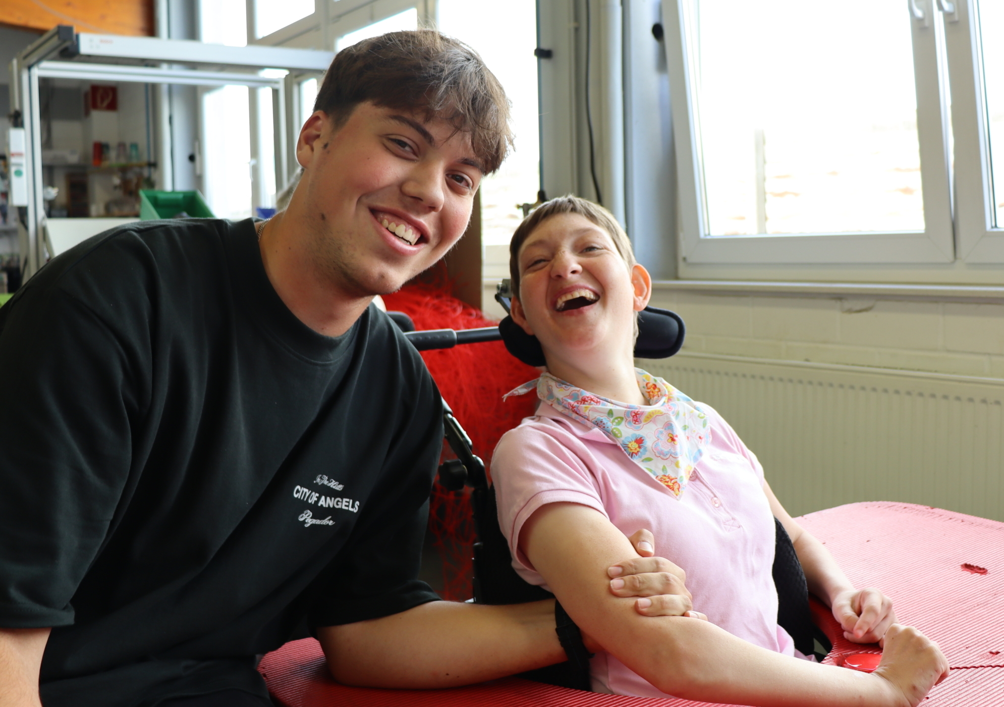 Ein Junge und ein Mädchen sitzen auf Stühlen. Beide lächeln. Das Mädchen trägt ein rosa Shirt und eine Krawatte.