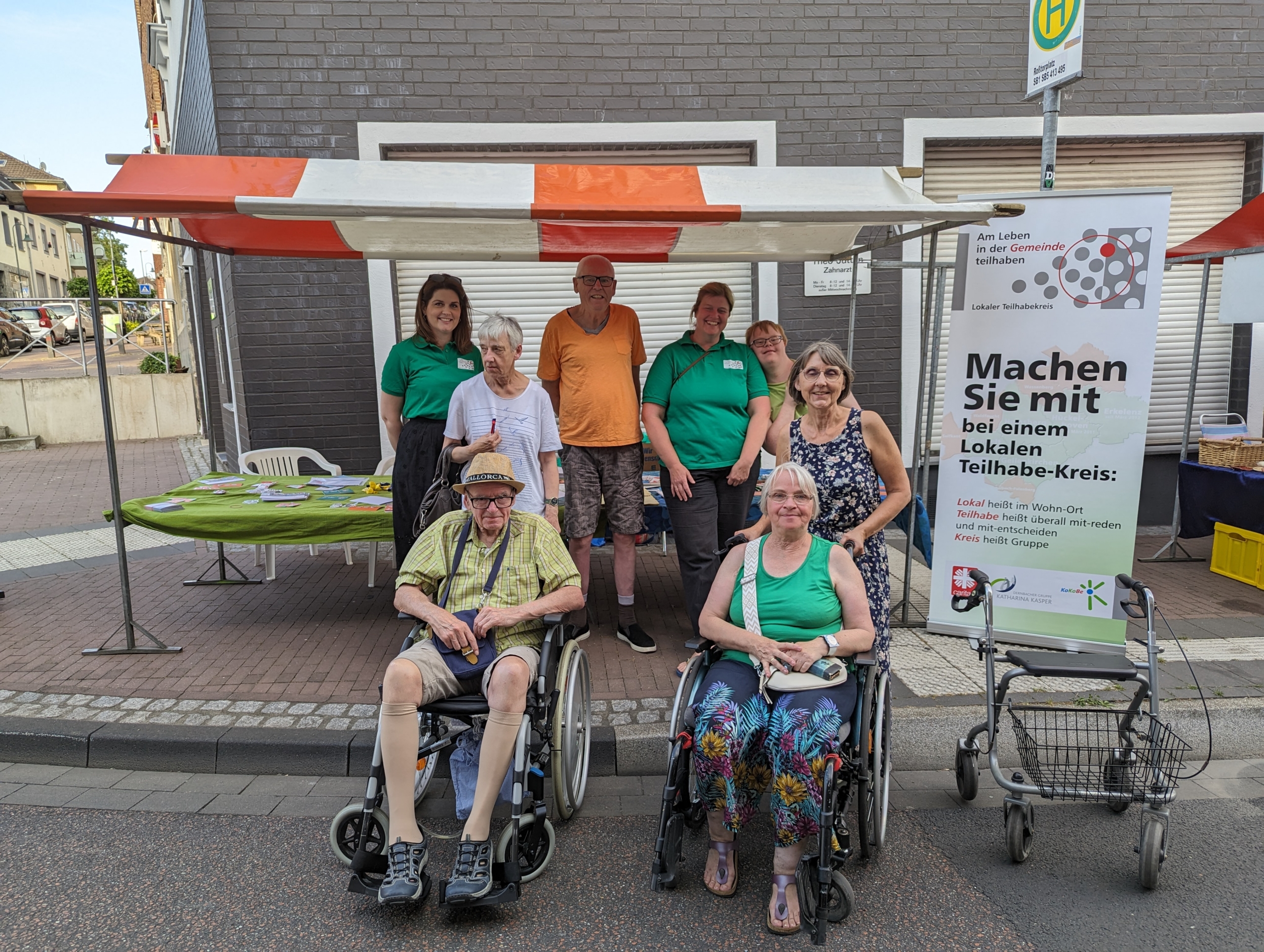 Gruppe von Menschen in Rollstühlen und Stehenden vor einem Informationsstand, der auf Barrierefreiheit und Inklusion hinweist.