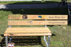 Eine Bank steht auf einem grünen Rasen. Es sind Bäume im Hintergrund zu sehen.
