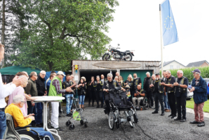Gruppenszene bei einem Motorradtreffen, wo viele Teilnehmer in Lederjacken versammelt sind, umgeben von Bäumen und einem Stand im Hintergrund.