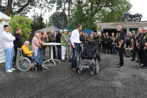Gruppe von Menschen, die sich im Freien versammelt haben, darunter ein Mann mit einem Kinderwagen, während andere an einem Tisch stehen. Die Szene findet in einer ländlichen Umgebung mit Bäumen und einem Holzschuppen im Hintergrund statt.