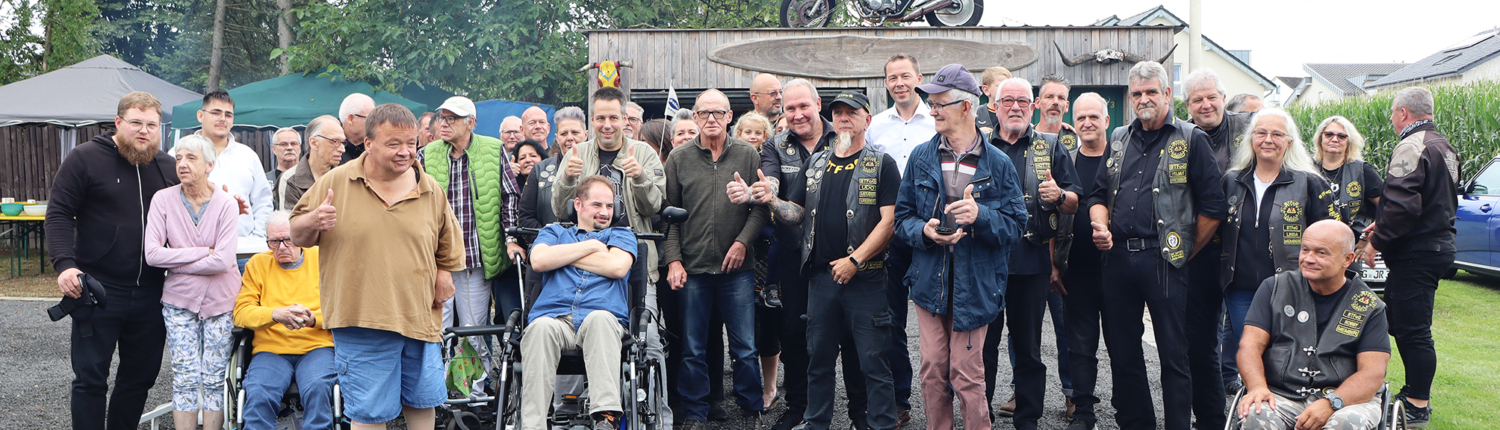Eine Gruppe von Menschen steht vor einer Hütte. Einige sitzen im Rollstuhl. Es sind viele Männer mit Motorrad-Jacken.