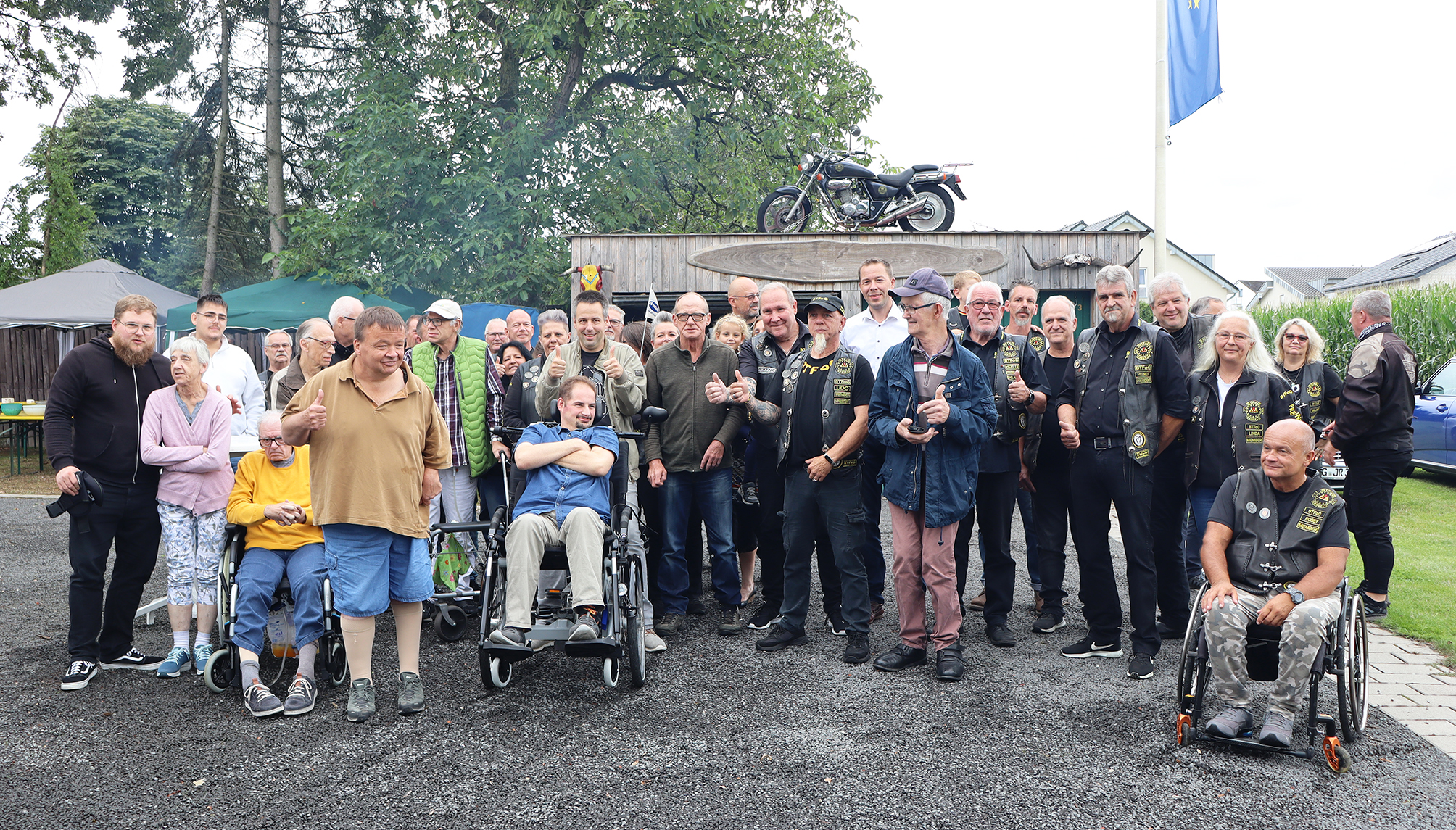 Eine Gruppe von Menschen steht vor einer Hütte. Einige sitzen im Rollstuhl. Es sind viele Männer mit Motorrad-Jacken.