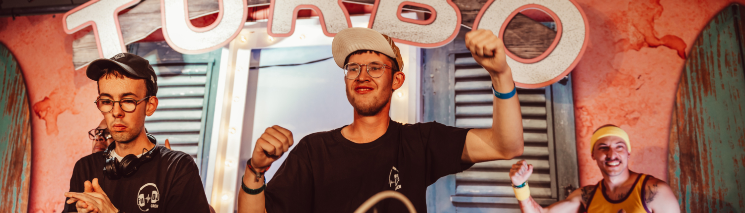Drei Männer stehen hinter einem DJ-Pult. Zwei von ihnen schauen in die Kamera, einer ist abgewandt.