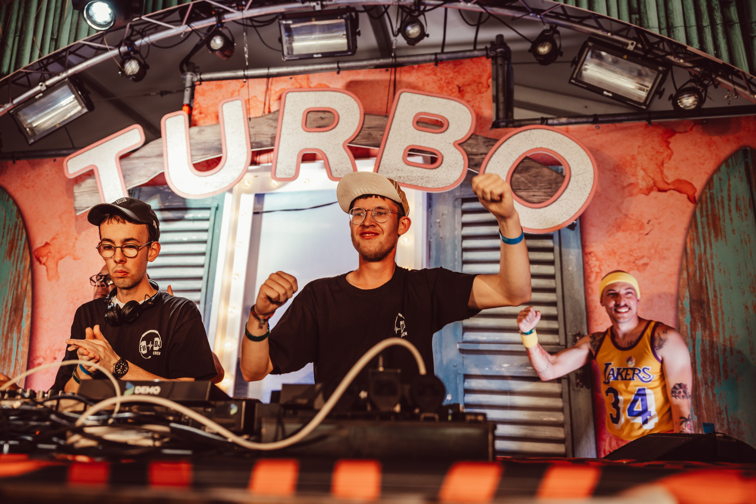 Drei Männer stehen hinter einem DJ-Pult. Zwei von ihnen schauen in die Kamera, einer ist abgewandt.