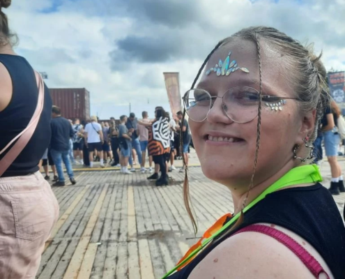 Junge Frau mit Brille und buntem Outfit, lächelt und sitzt auf einer Holzplattform, während im Hintergrund eine Menschenmenge bei einem Event oder Festival zu sehen ist.