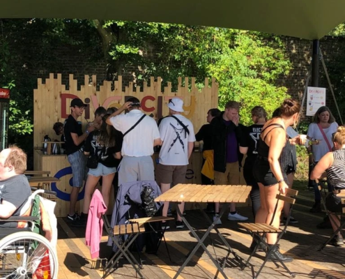 Menschen stehen an einem Stand im Freien, einige sind im Rollstuhl, während andere an Tischen sitzen und sich unterhalten. Die Szenerie zeigt eine gesellige Atmosphäre bei einem Event oder Festival.