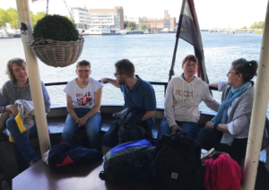 Leute sitzen auf einem Boot. Man sieht Wasser und Gebäude im Hintergrund.