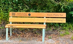 Eine Bank steht im Wald. Die Bank hat Holz und Metall. Es sind Bäume im Hintergrund.