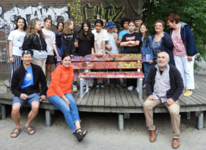 Gruppe von Jugendlichen und Erwachsenen, die gemeinsam auf einer Holzbank sitzen und stehen, umgeben von Grünanlagen, bei einer Freizeitaktivität oder einem Event.