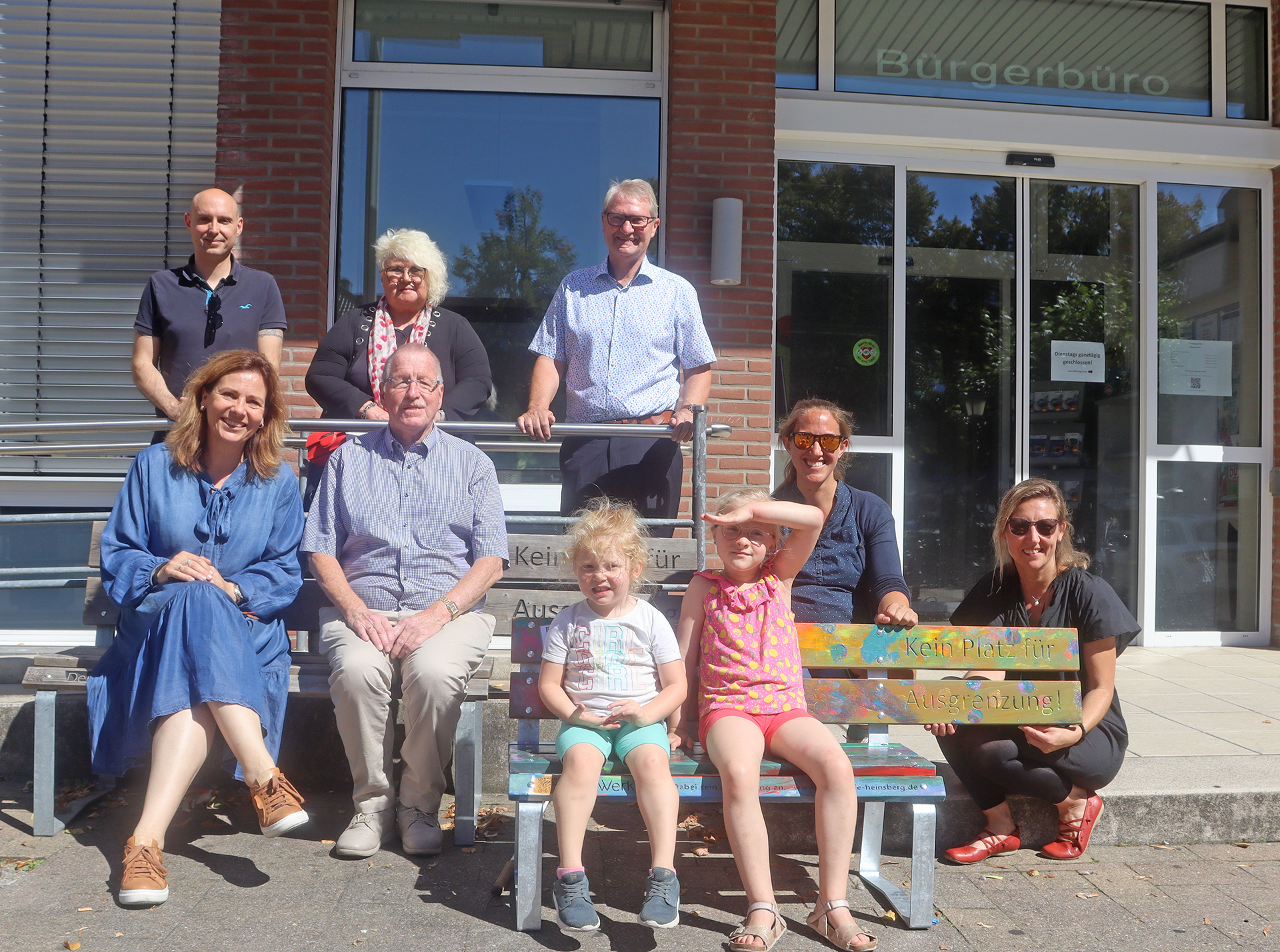 Menschen sitzen auf einer Bank vor einem Gebäude. Ein Kind steht auf der Bank.